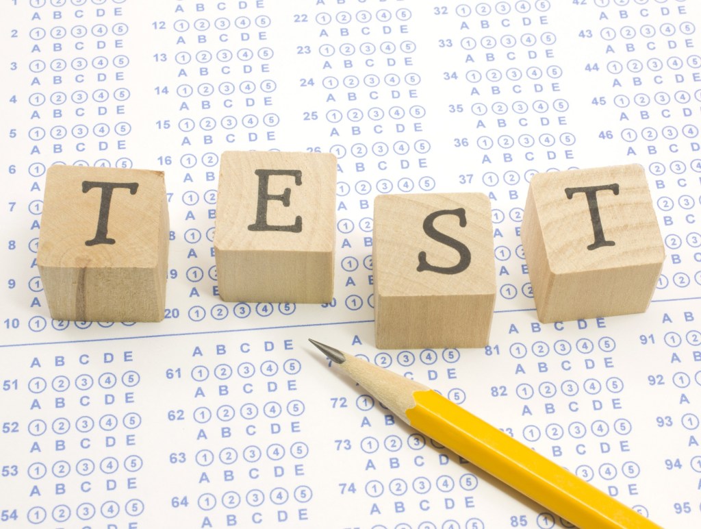 Wooden blocks spell out TEST on a blue bubble or scantron sheet with a number two yellow pencil.